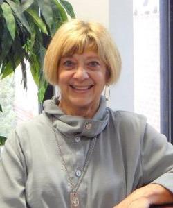 Smiling woman with short blonde hair, 穿着霍华德·佩恩大学的灰色外套，戴着项链, seated indoors. | HPU